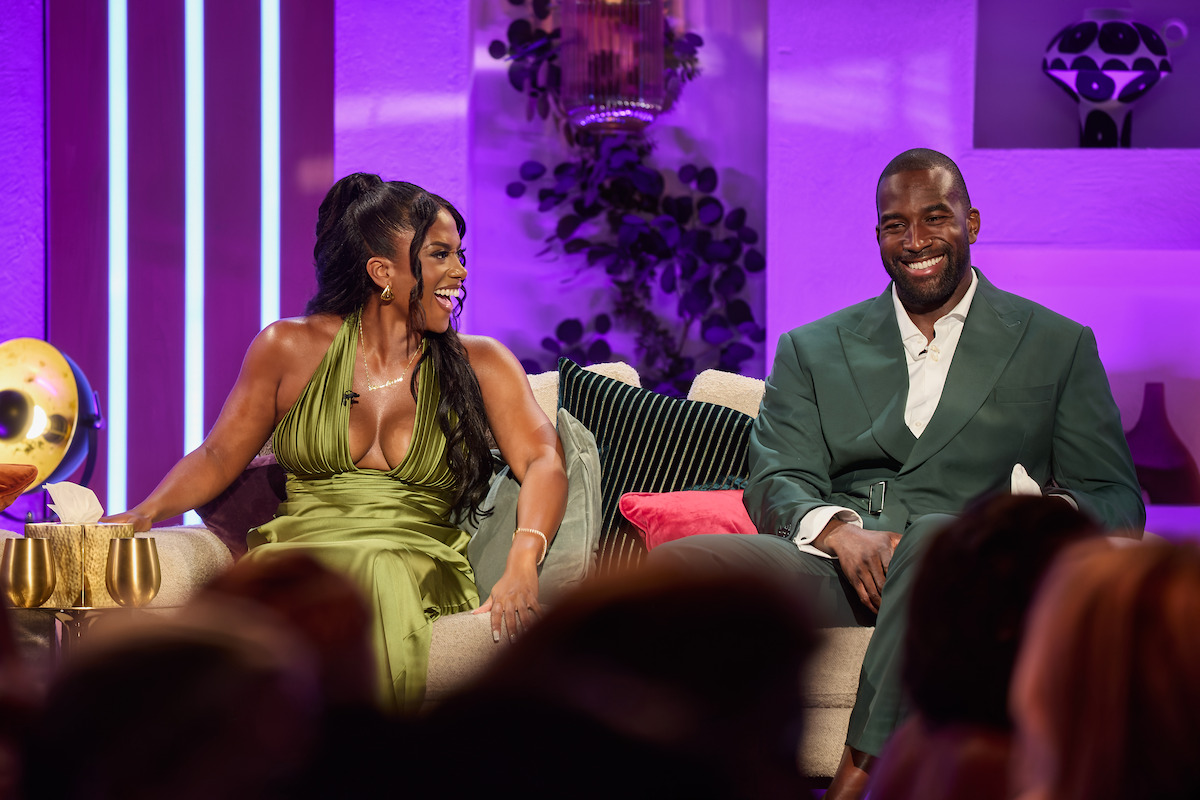 Two people in formal attire sit on a modern sofa in a brightly lit studio with pink and purple lighting, decorative plants, and contemporary decor, smiling and engaging with the audience.