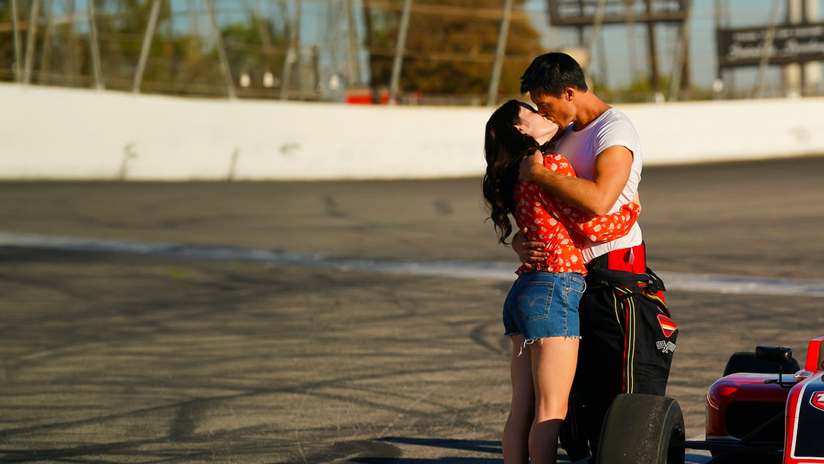 A couple kisses on a racetrack beside a red race car, with the driver in racing attire and the woman in casual clothes, sunlight illuminating the scene.