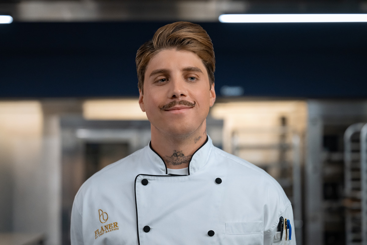 A confident male chef in a white uniform stands in a commercial kitchen, with stainless steel appliances and racks visible in the background.