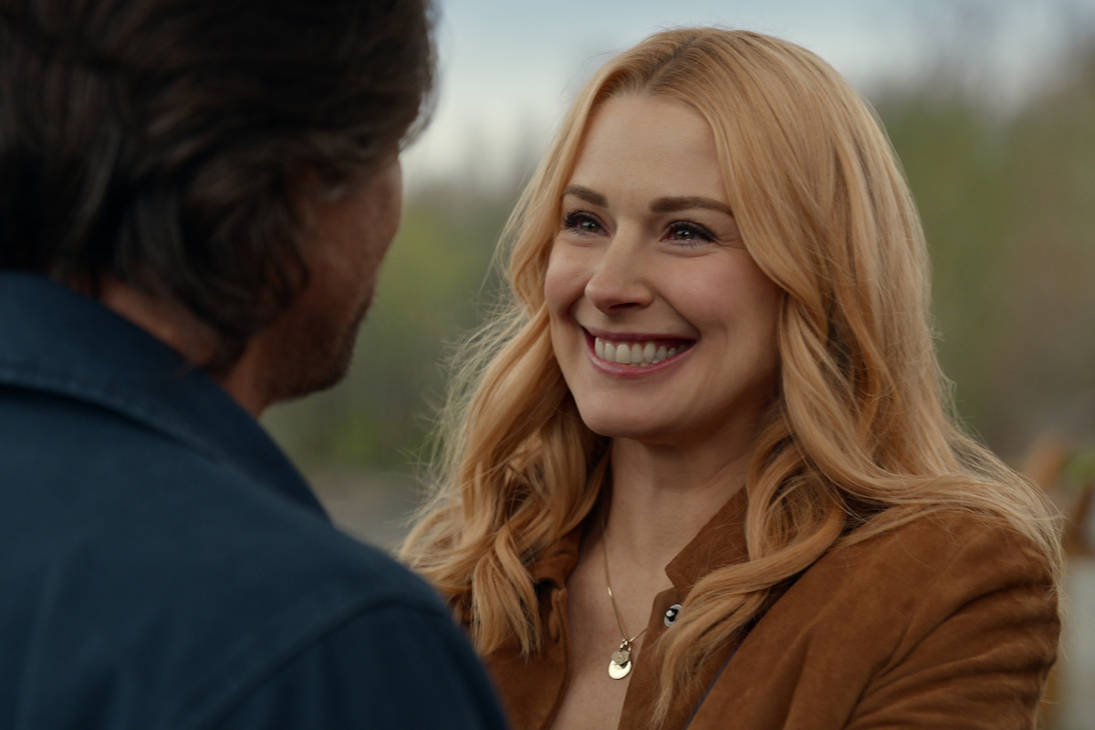 A blonde woman smiles warmly at a man outdoors, both wearing jackets. The background is blurred with natural greenery, suggesting a happy, intimate moment in a peaceful, rustic setting.
