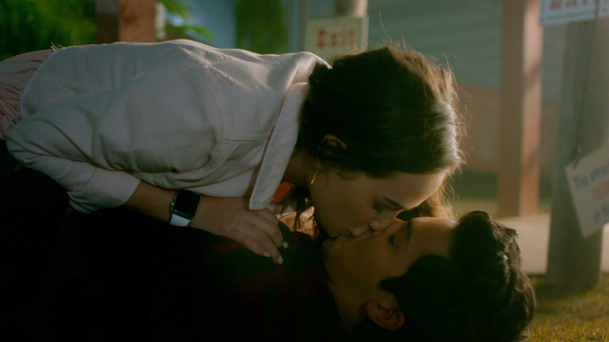 A young woman kisses a young man while lying on the grass at night, with soft light and outdoor surroundings, including signs and greenery in the background.