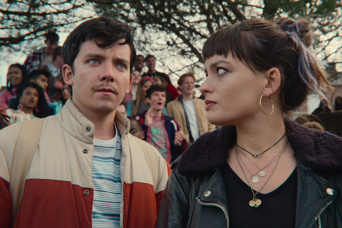 Asa Butterfield as Otis Milburn and Emma Mackey as Maeve Wiley stand together outside with a crow behind them in Season 3 of ‘Sex Education.’