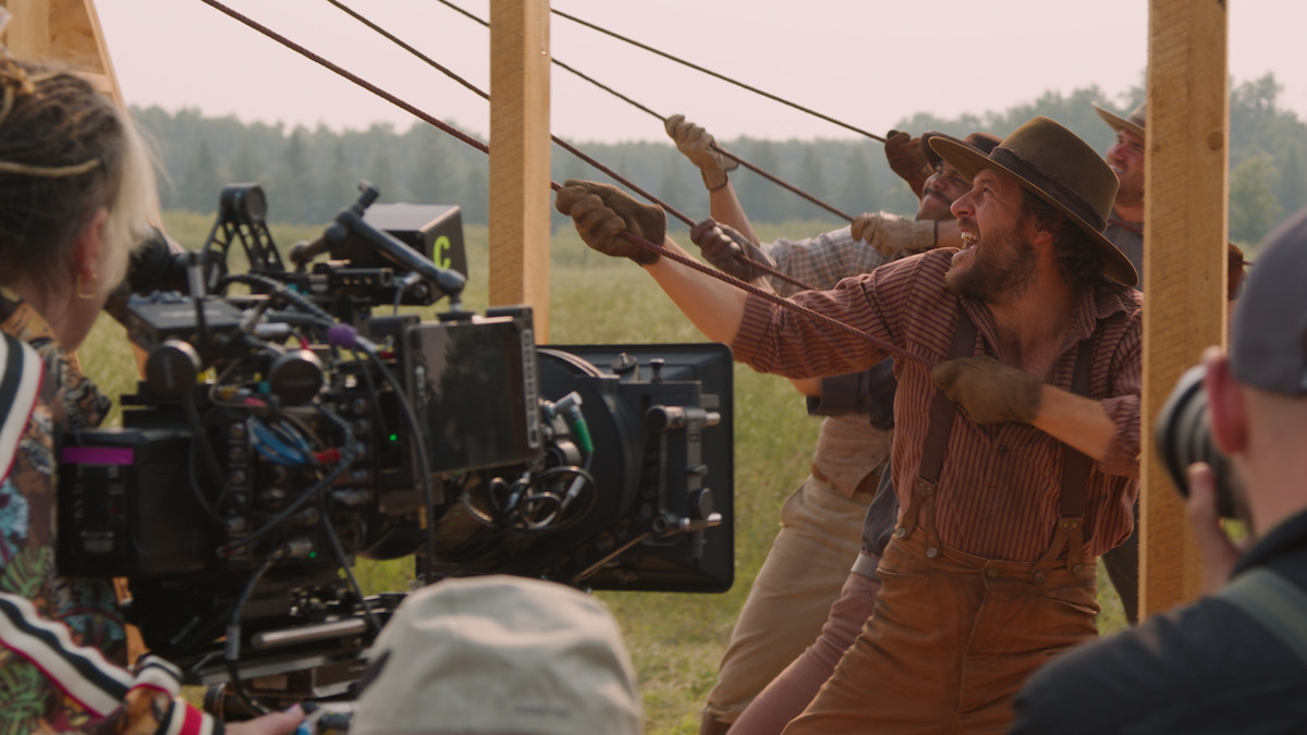 Film crew shoots a scene outdoors with actors in period costumes pulling ropes on a wooden structure; camera and crew visible in foreground, grassy field and trees in background.