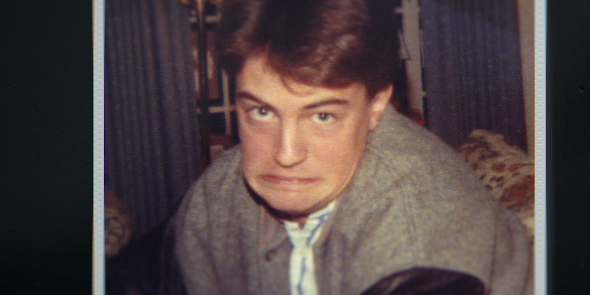 Young man indoors making a humorous, exaggerated face, wearing a grey jacket with leather sleeves. The environment looks like a living room with curtains and patterned furniture in the background. Warm, casual setting.