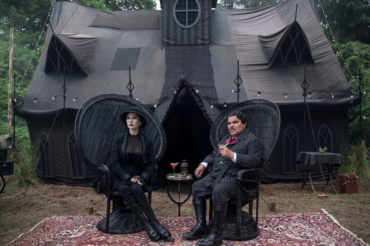 Catherine Zeta-Jones como Morticia Addams y Luis Guzmán como Gómez Addams se sientan en sillas negras frente a una tienda negra en la temporada 2 del 'Miércoles'.
