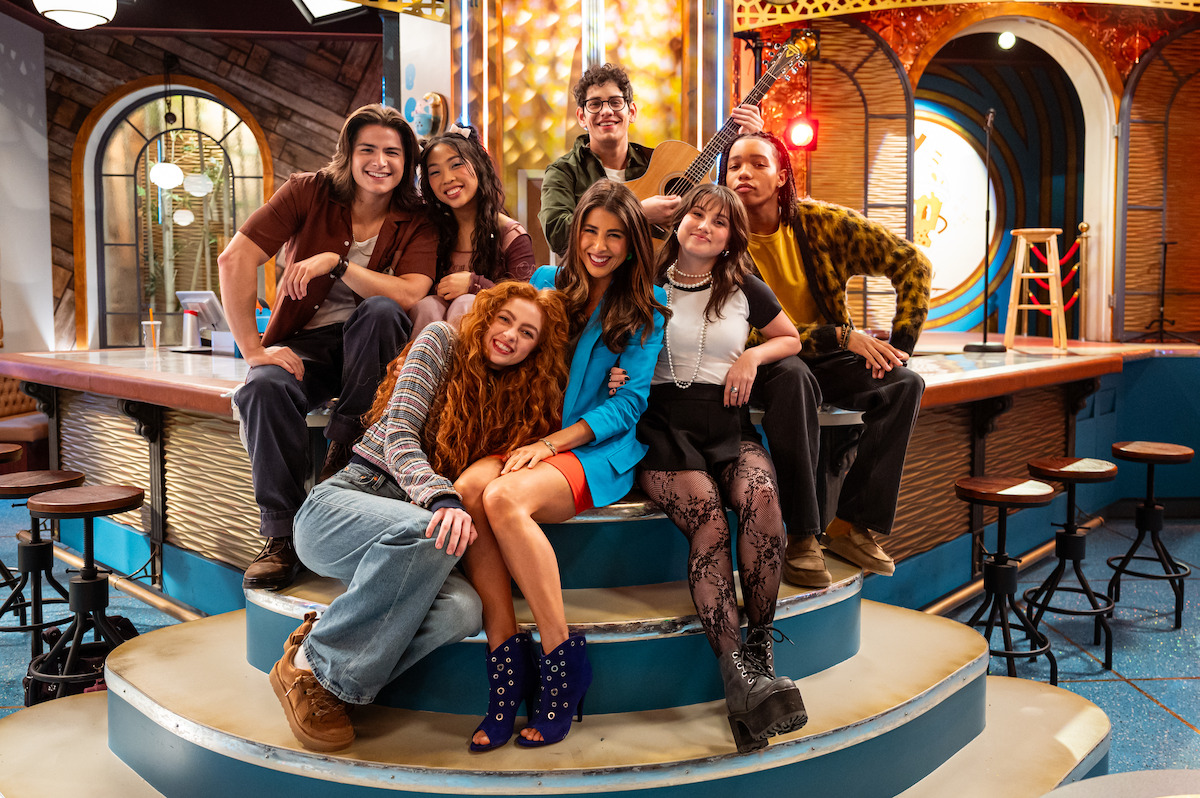 Seven young adults pose together smiling inside a colorful, modern, well-lit café with barstools, arched windows, decorative walls, and a stage in the background. One person holds a guitar, capturing a cheerful and lively atmosphere.