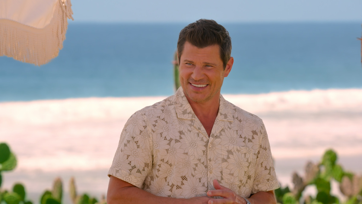 Man in a floral shirt smiling on a sunny beach, with ocean waves and greenery in the background, under a light-colored umbrella.