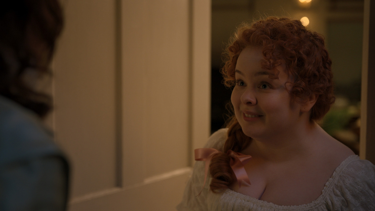 A young woman with curly red hair and a ribbon smiles warmly in a softly lit room, wearing historical clothing. She stands near a doorway, facing someone just out of the frame, with warm light in the background.