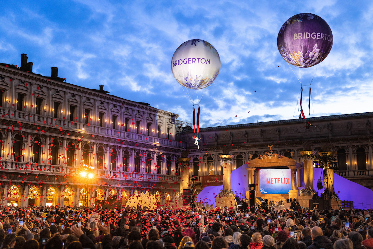 Grande multidão se reúne em uma praça histórica ao anoitecer para um animado evento de ‘Bridgerton’, da Netflix, com holofotes, decorações e dois grandes balões de hélio iluminados exibindo o logotipo da série no céu, logo acima de edifícios ornamentados.