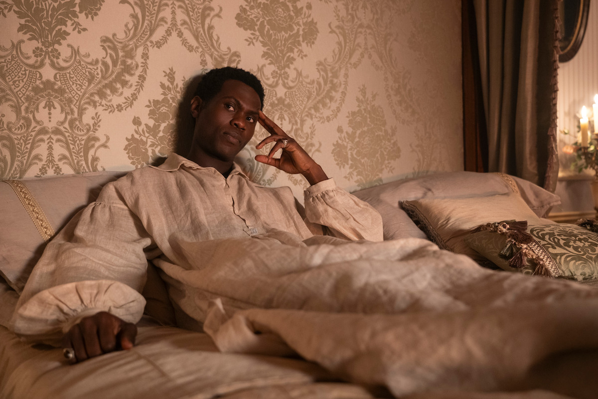 A man in historical nightwear sits up in an ornate bed, resting his head on his hand. The room has elegant wallpaper, decorative pillows, and soft candlelight, giving a vintage, luxurious atmosphere.