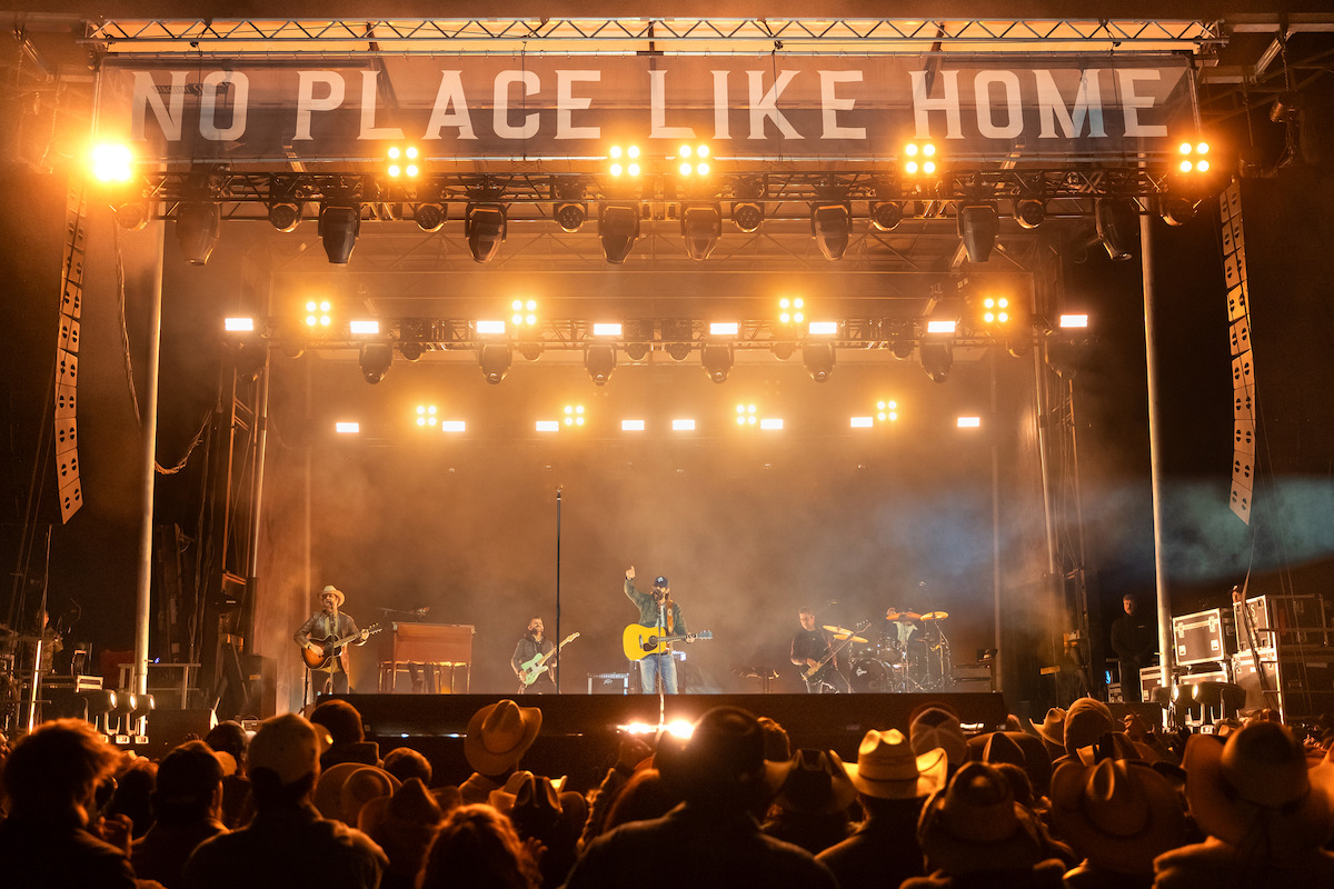 A band performs on a brightly lit outdoor stage at night with "NO PLACE LIKE HOME" displayed above, while a crowd in hats watches the concert.