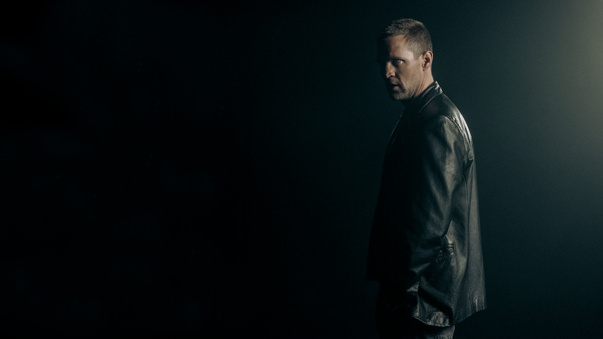 A man in a leather jacket stands in a dark, moody environment with dramatic lighting from behind, creating a tense and mysterious atmosphere.