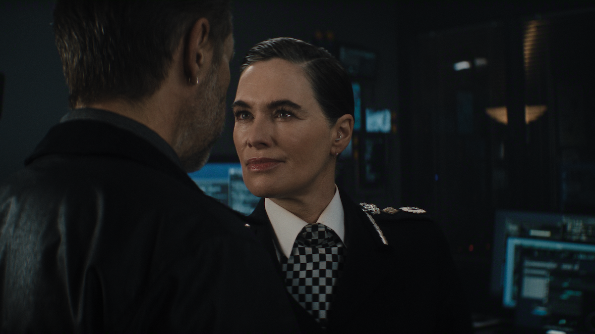 A stern police officer in uniform faces a man in a dimly lit control room, surrounded by computer monitors and electronic equipment, creating a tense atmosphere.