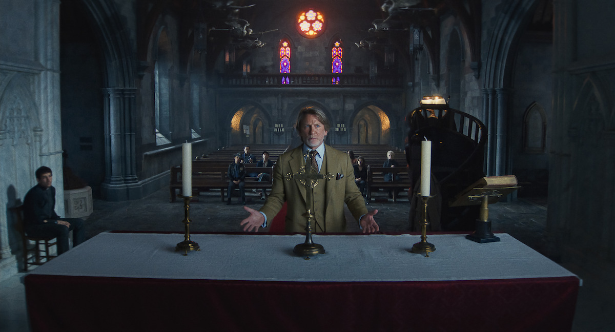 A man in a suit stands behind an altar in a dimly lit, Gothic church with stained glass windows. Several people sit in pews, creating a solemn and tense mood. The environment is grand, mysterious, and atmospheric.