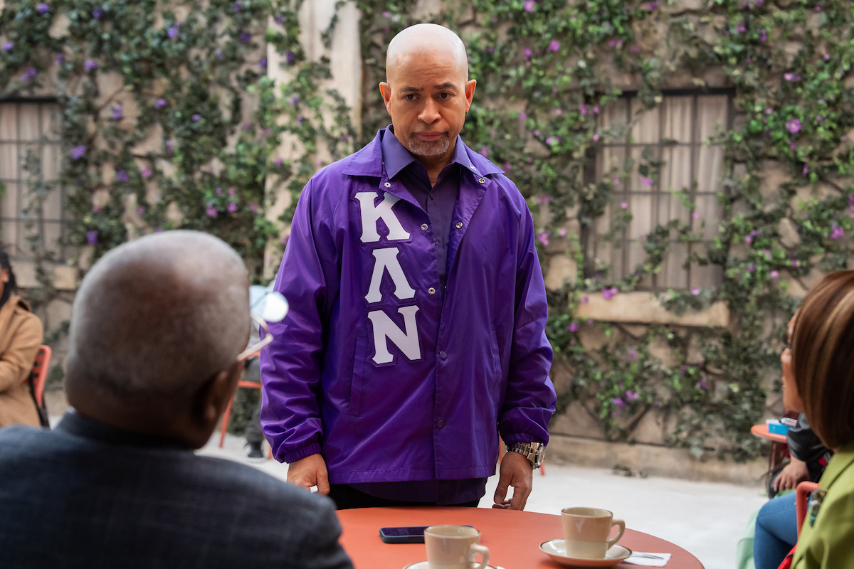 A man in a purple jacket with white letters stands by a table with cups, facing two seated people outdoors. Greenery and flowers cover the wall behind them, suggesting a relaxed café or patio setting.