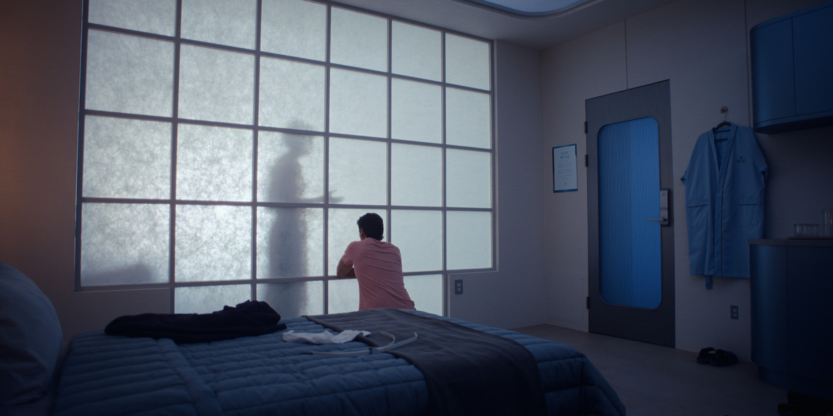 A person kneels by a frosted glass wall with a shadowy figure behind it in a sterile, modern room with a neatly made bed, blue linens, a robe on the wall, and medical or institutional furnishings.