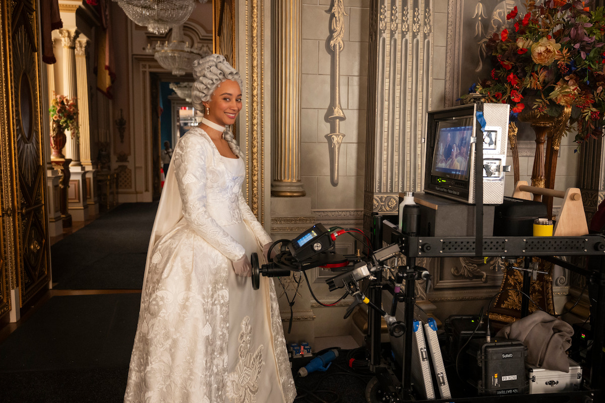 Woman in elaborate white historical gown and wig stands on ornate film set next to video equipment and monitor, surrounded by decorative walls and flowers, smiling towards the camera.