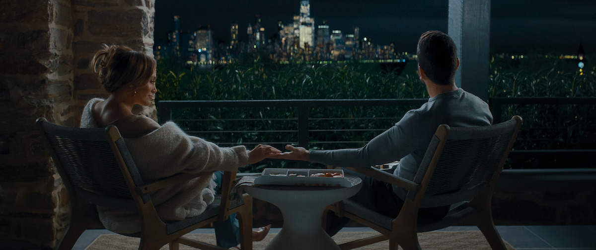 Two people sit on a balcony at night holding hands, facing a city skyline illuminated in the distance, with a small table and cozy seating, suggesting a peaceful, intimate setting.