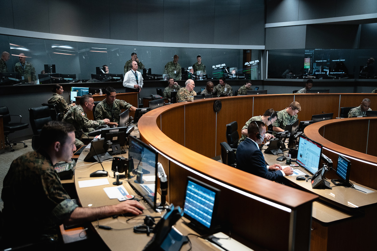 Military personnel and officials work at computer stations in a modern command center with curved desks, large monitors, and dim lighting, suggesting a serious, focused mood and high-security environment.