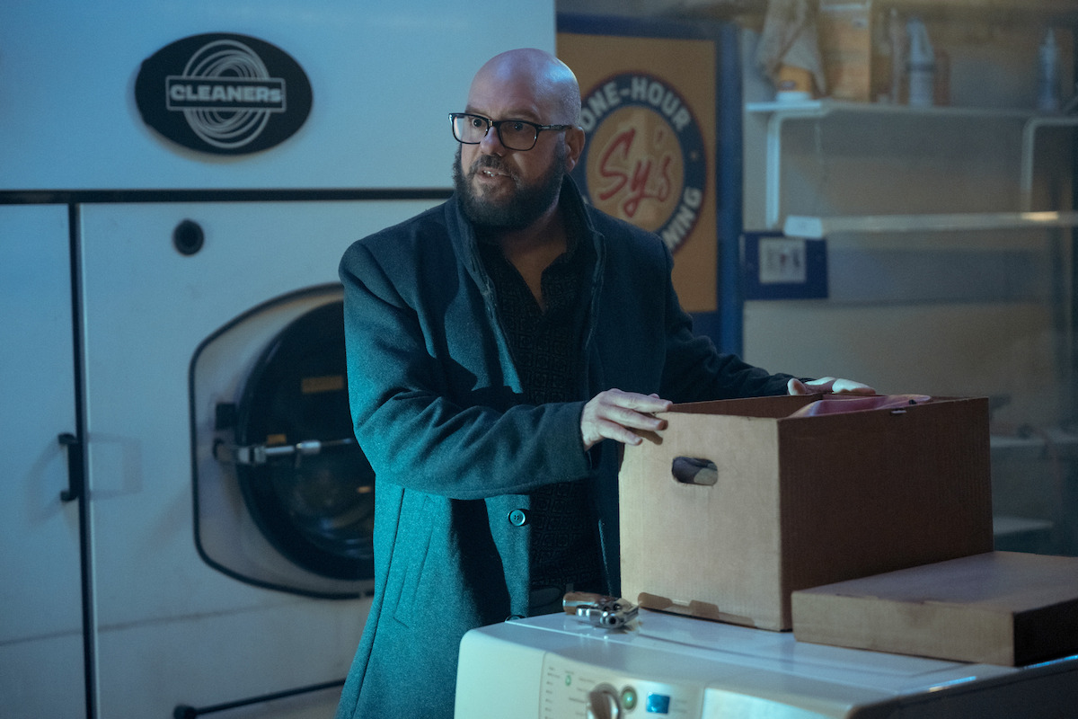 David Cross as Sy Grossman stands in a dry cleaning shop holding a brown box in Season 4 of ‘The Umbrella Academy’