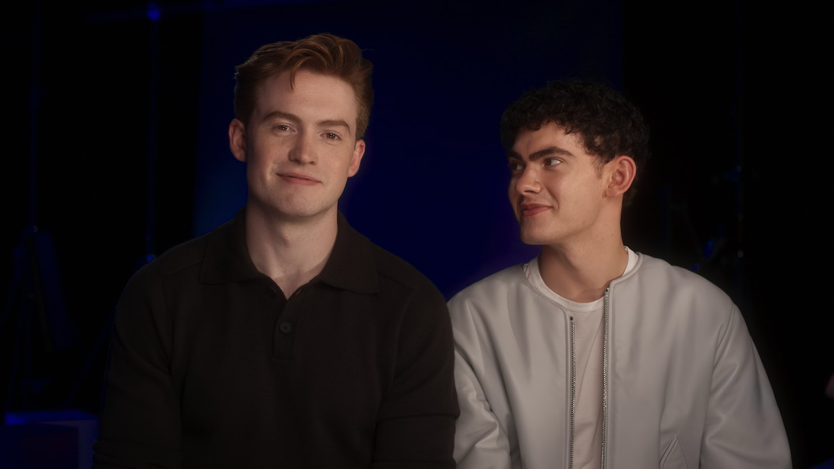Two young men sitting closely together in a dimly lit indoor setting, one in a dark shirt smiling calmly at the camera while the other, in a light jacket, looks at him with a fond expression.
