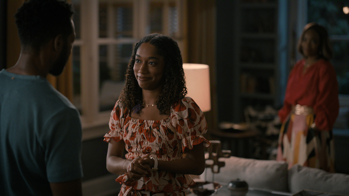 A young woman smiles and talks to a man in a warmly lit living room, with another woman standing in the background near a window and bookshelf. The setting appears cozy and intimate, suggesting a personal or emotional conversation.