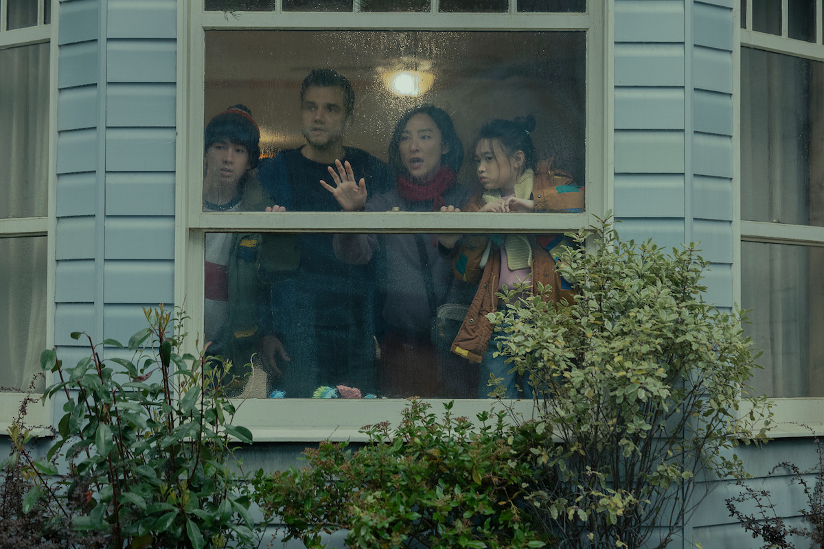 Four people stand inside a house looking out a rain-covered window with concerned expressions; the scene is set in a cozy, well-lit room with plants outside the window.
