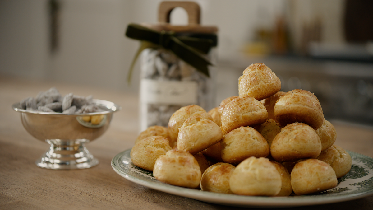 A plate of golden, baked pastry puffs on a decorated dish, with a bowl of powdered treats and a jar with a ribbon in a cozy, softly lit kitchen setting, evoking a warm, inviting mood.