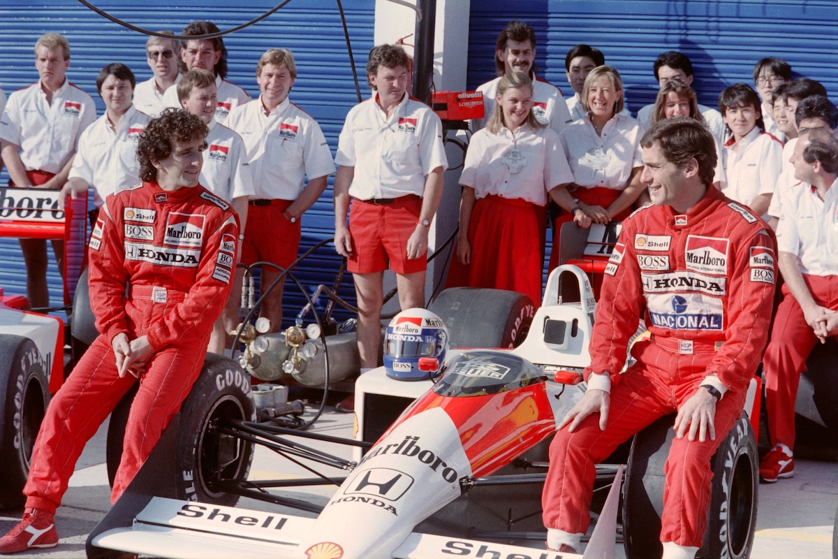 Alain Prost and Ayrton Senna sit on the front wheels of their two red and white McLaren Formula 1 cars with their team standing behind them as they pose for photographers.