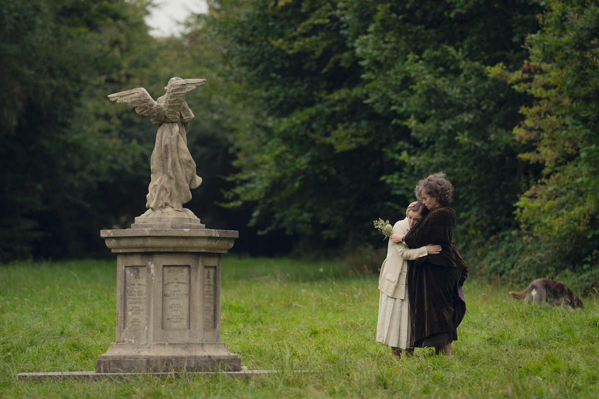 Two people embrace in a grassy clearing near a stone angel statue, surrounded by dense green trees. A dog is in the background. The setting feels calm and somber, resembling a countryside or park-like memorial.