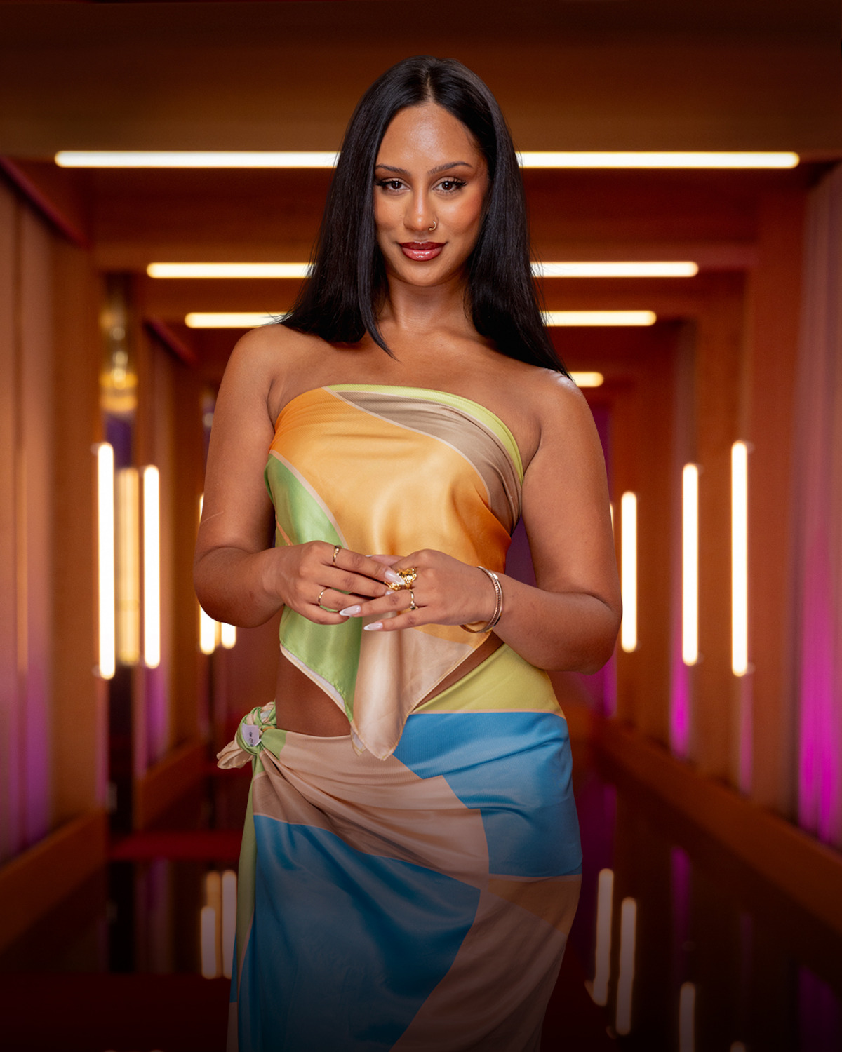 Woman with long dark hair wearing a colorful, strapless dress stands in a modern, warmly lit hallway with glowing vertical lights on the walls and soft pink accents in the background.