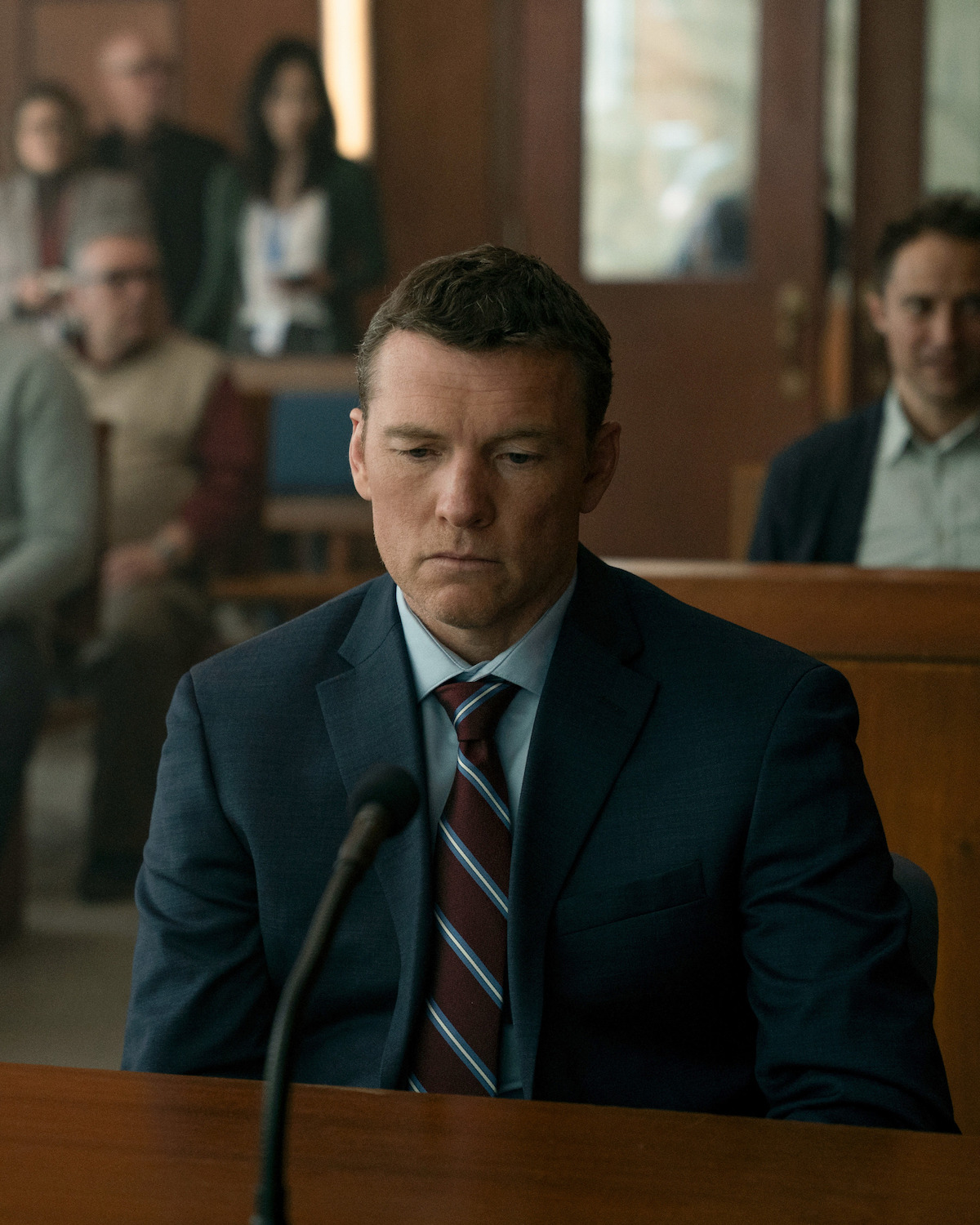 A man in a blue suit sits at a courtroom table in front of a microphone, looking serious and focused, with several people seated in the background observing the proceedings.