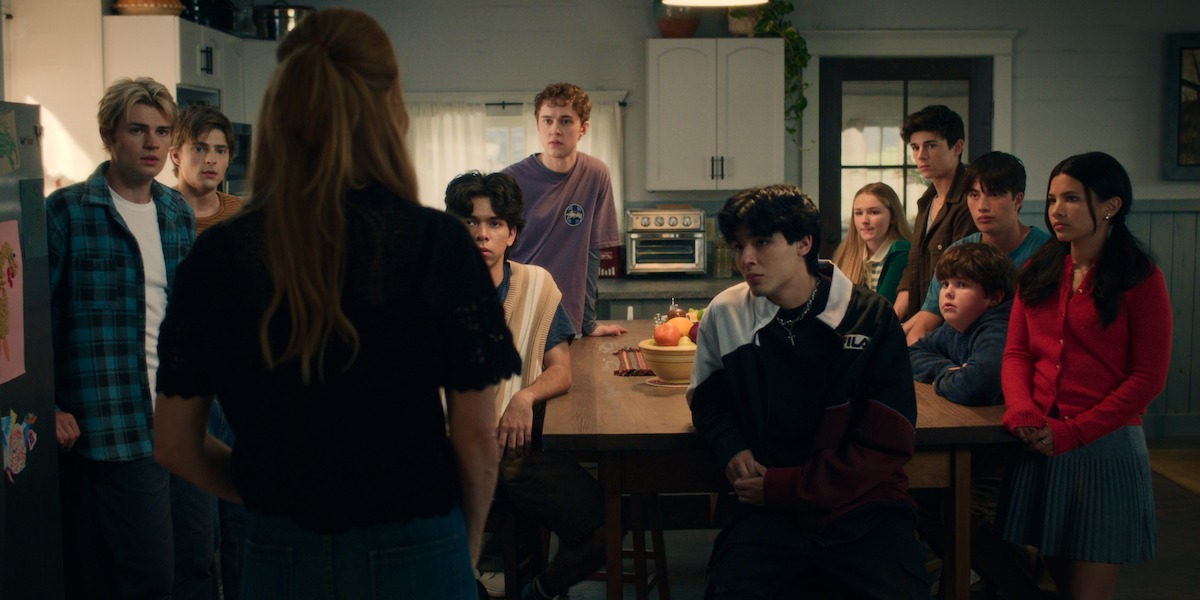 A woman stands facing a group of teenagers gathered around a kitchen table in a cozy, warmly lit home, all focused on her as if listening intently to an important conversation.
