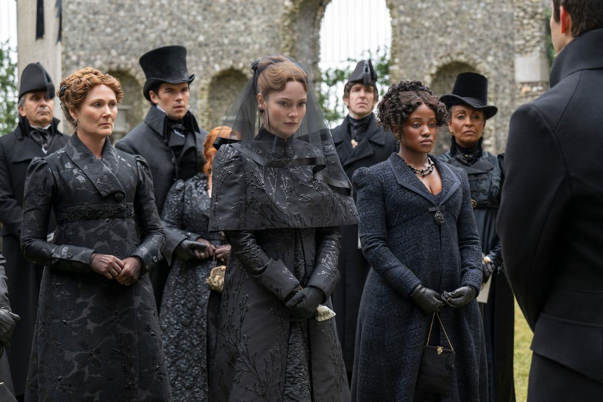 A group of people in dark, formal Victorian-era attire stand outdoors near stone ruins, appearing somber as if attending a funeral or serious gathering.