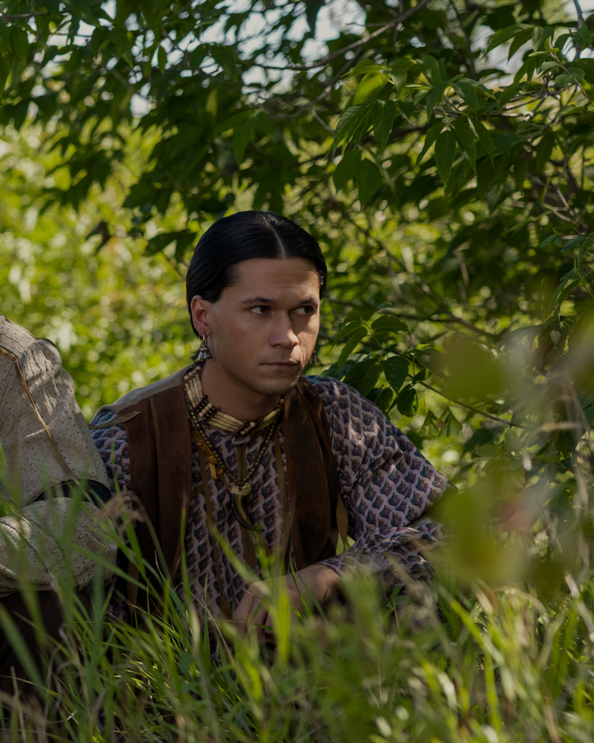 A young person with dark hair, wearing patterned clothing and a leather vest, sits among tall grass and green foliage in a natural outdoor setting, partially shaded by overhanging tree branches.