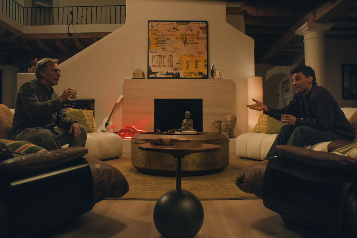 Two men are sitting on opposite sofas in a cozy living room having a conversation. The room has warm lighting, art and musical instruments by the fireplace, a round coffee table, armchairs, and decorative objects.