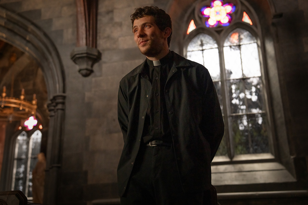 Young priest standing and smiling in a dimly lit, historic stone church with arched stained-glass windows, giving a warm and hopeful atmosphere.