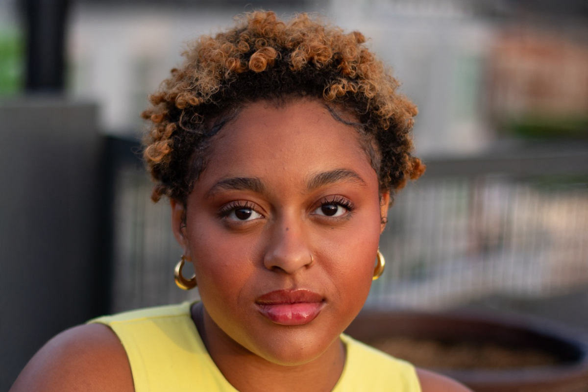 Person with short curly hair, gold earrings, and a yellow top poses outdoors in natural light, with a blurred urban background featuring a metal railing and buildings.