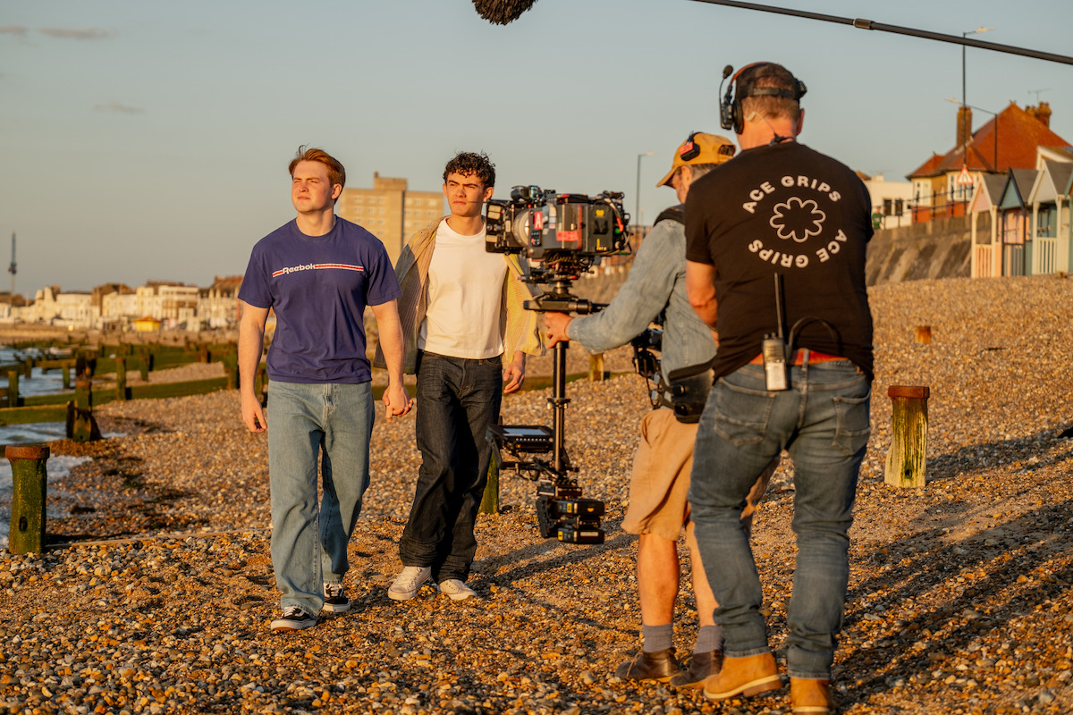 Two actors walking on a pebbled beach during sunset, being filmed by a crew with professional camera and boom mic; colorful houses and seaside town in the background.