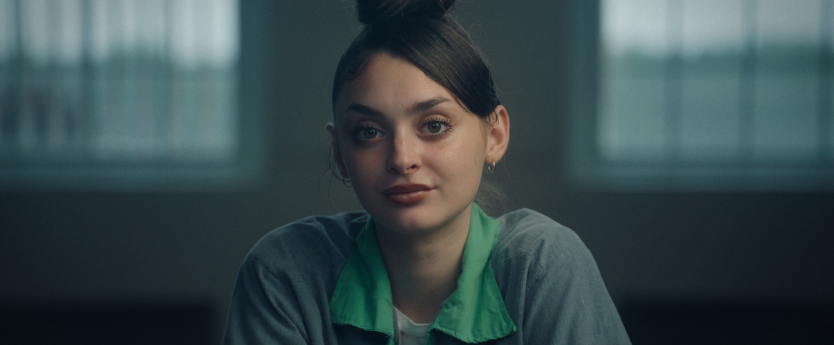 Young woman with dark hair in a bun sits indoors, wearing a grey and green collared shirt. She appears to be in a serious environment with barred windows, possibly an institutional setting like a prison or interrogation room.