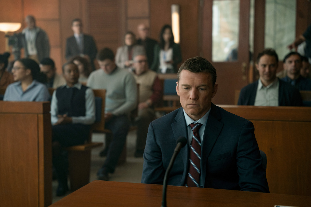 A man in a suit sits alone at a defendant's table in a courtroom, looking serious. Behind him, an audience of various people watches attentively from the wooden benches, suggesting a tense legal proceeding.