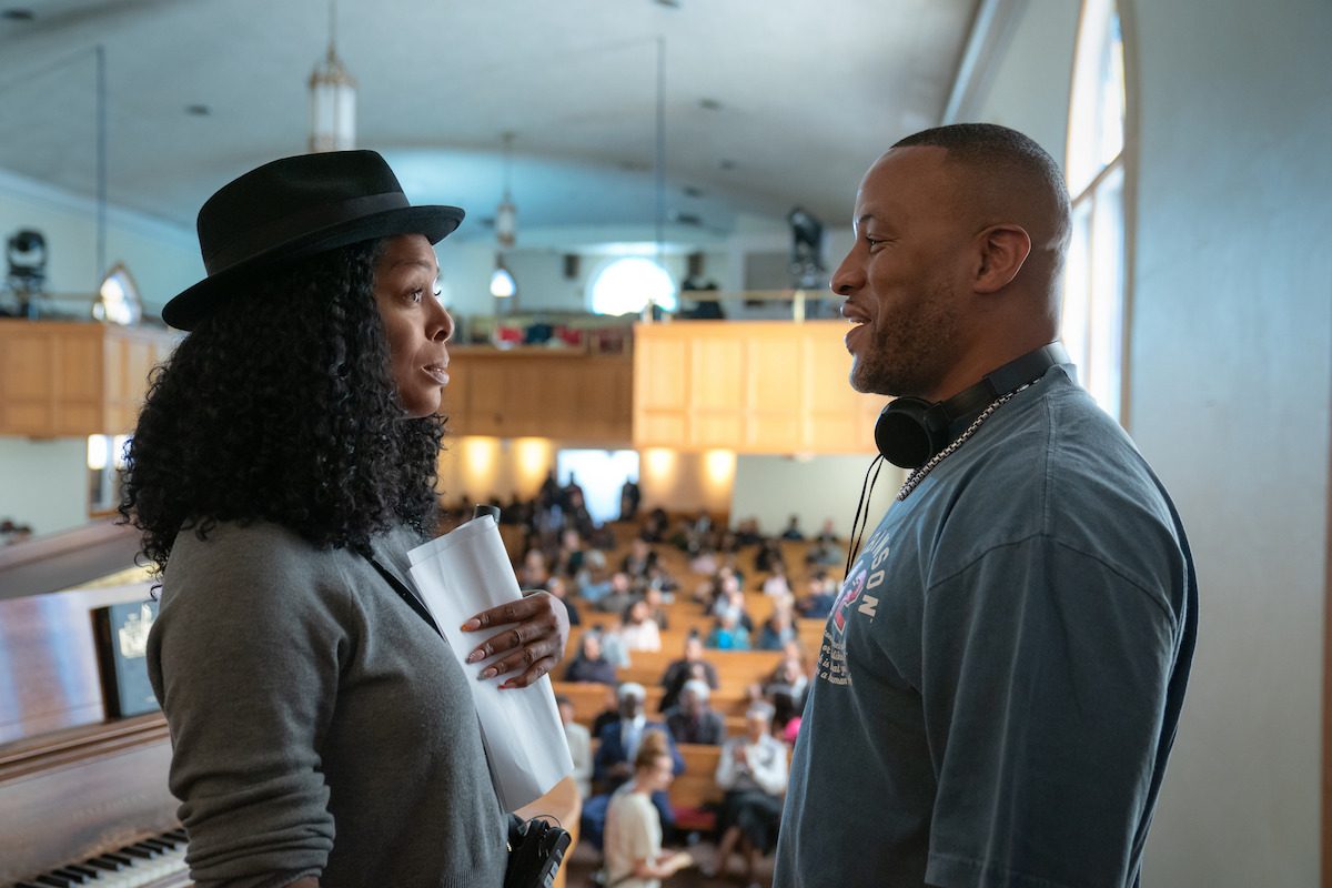 Two people have a conversation in a church or auditorium filled with people. One holds papers, the other wears headphones around his neck. Sunlight streams through windows, creating a warm, lively atmosphere.