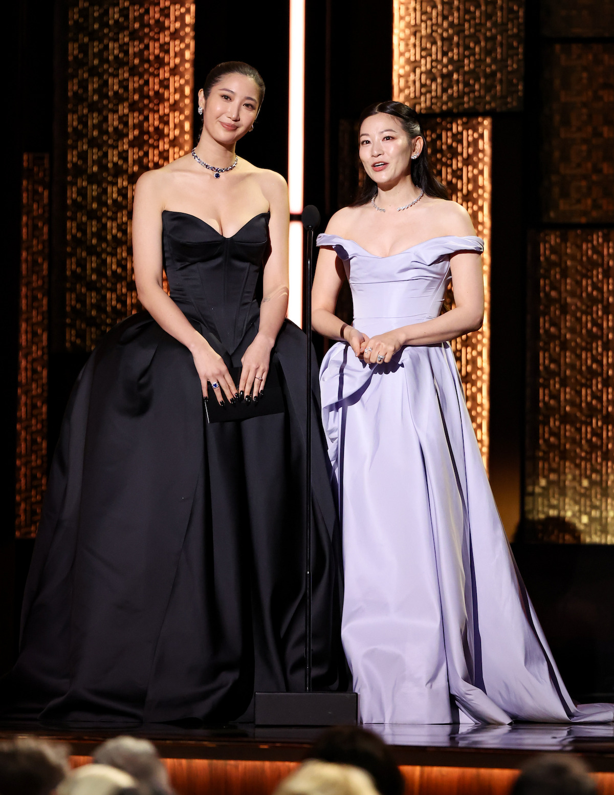 Two women in elegant evening gowns stand on stage in front of a microphone, presenting or speaking at a formal event with an illuminated decorative background.