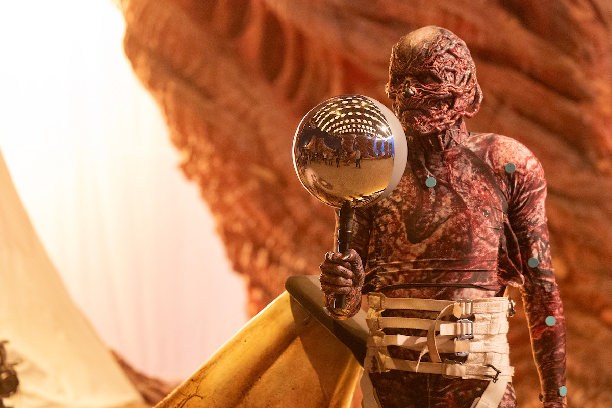 Actor in grotesque creature costume holds a reflective silver sphere on a stick against an organic-looking set backdrop.