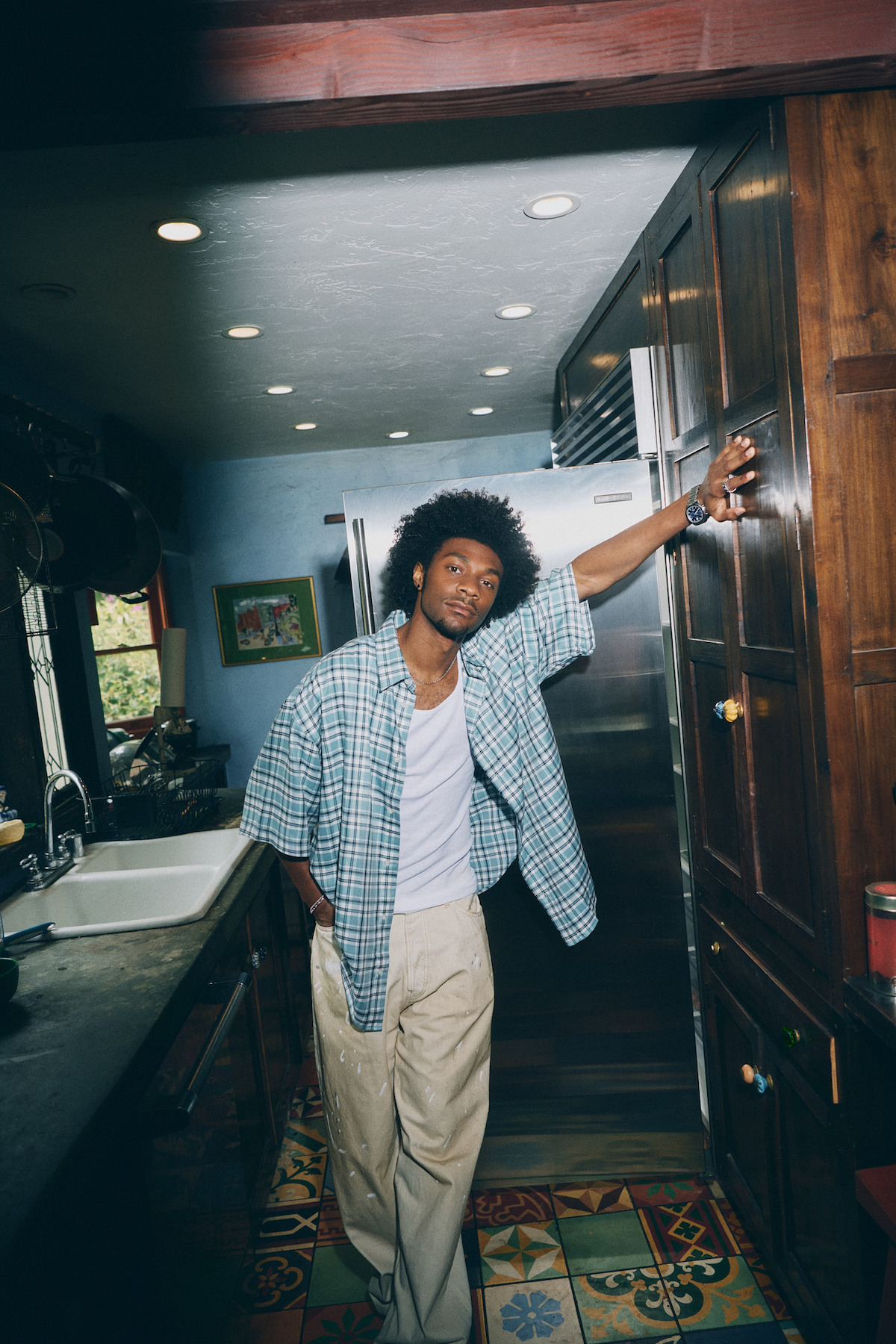 A person wearing a plaid shirt and light pants leans casually in a cozy kitchen with wooden cabinets, colorful tiled floor, and some wall art in the background. Indoor natural light and a relaxed atmosphere.