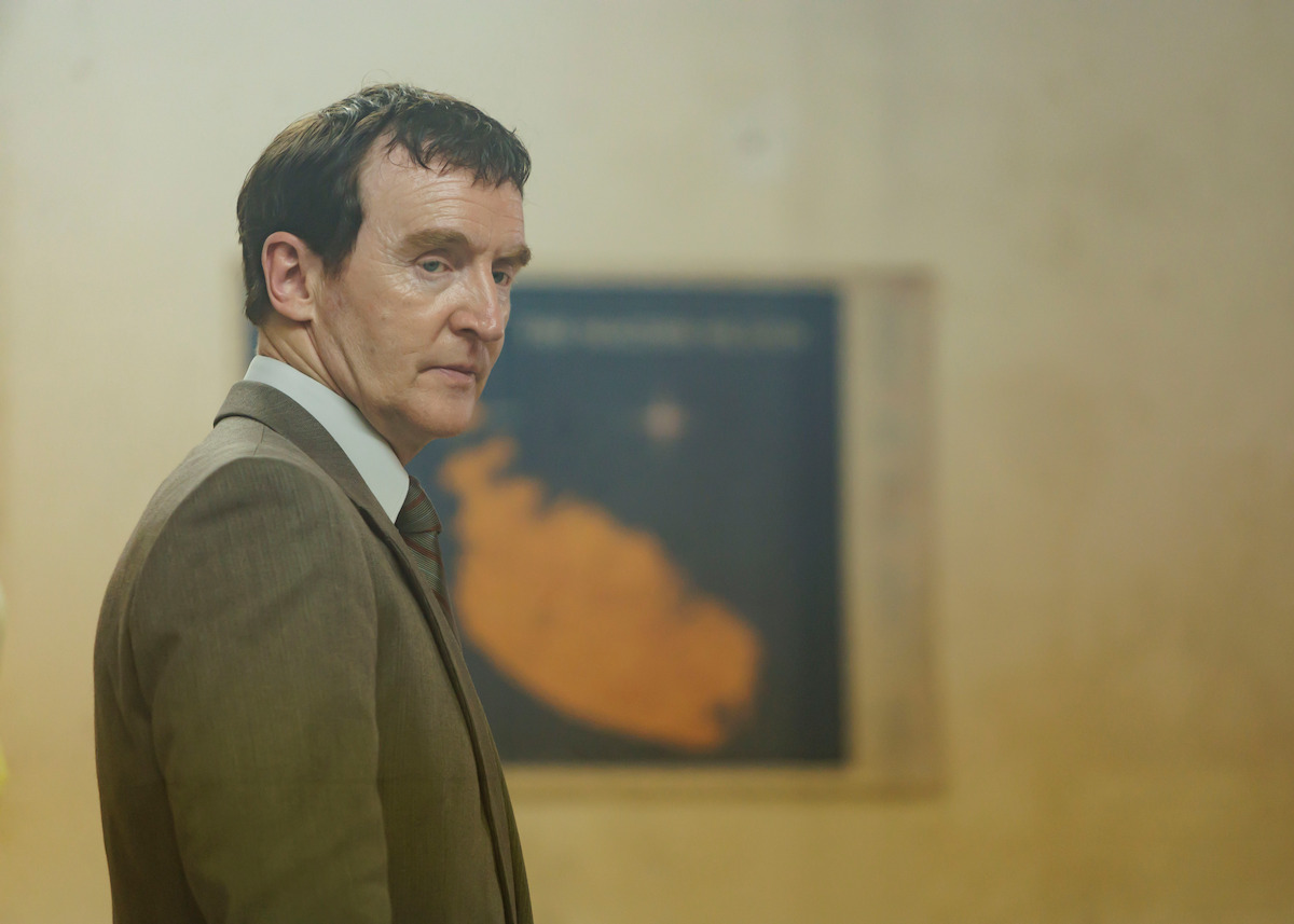 Man in a brown suit looking to the side, standing indoors in front of a blurred map or artwork on a beige wall, under soft lighting.