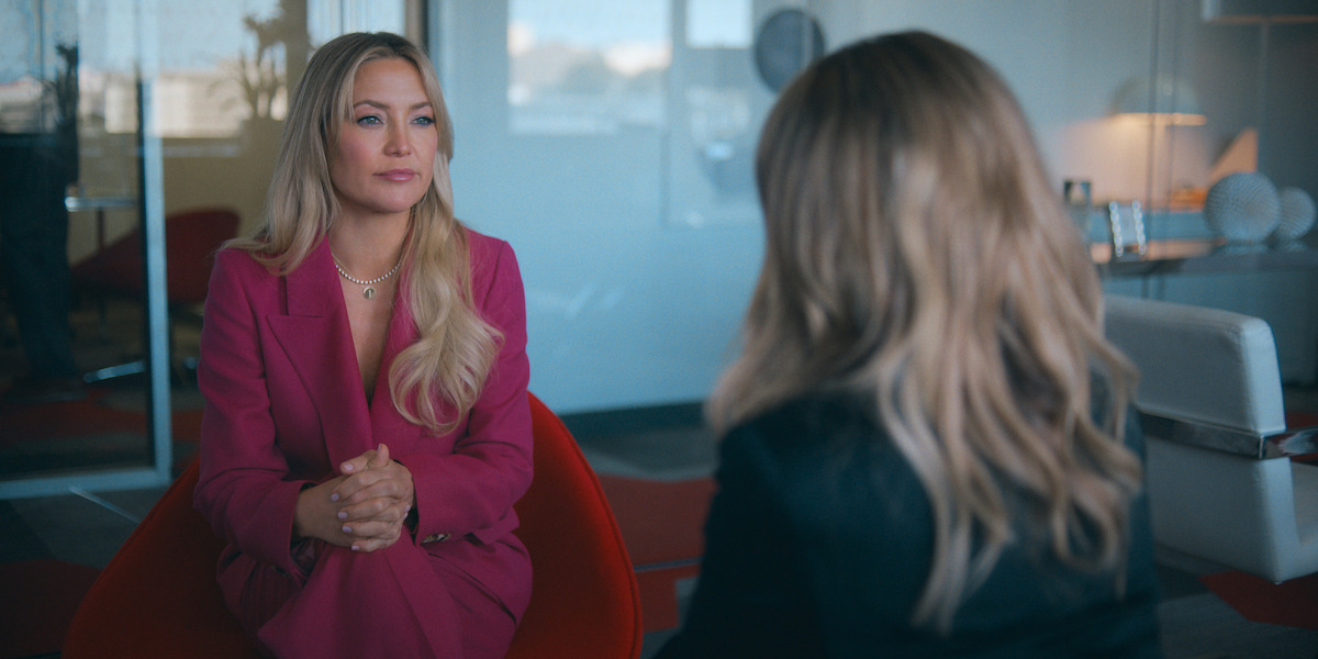 Two women sit facing each other in a modern office. One, in a pink suit, sits on a red chair with hands clasped, while the other, seen from behind, sits opposite. Glass walls and office furniture are in the background.