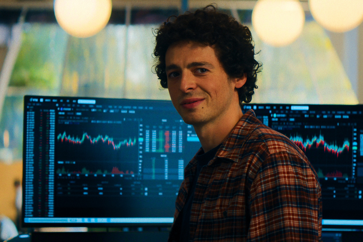 A man in a plaid shirt sits in front of computer monitors displaying financial charts and stock data in a modern office with large windows and warm lighting.