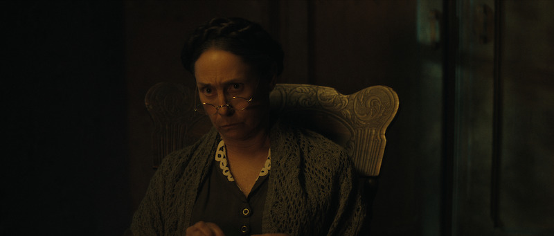 An older woman with glasses and braided hair sits in a carved wooden chair, looking sternly ahead in a dimly lit room.