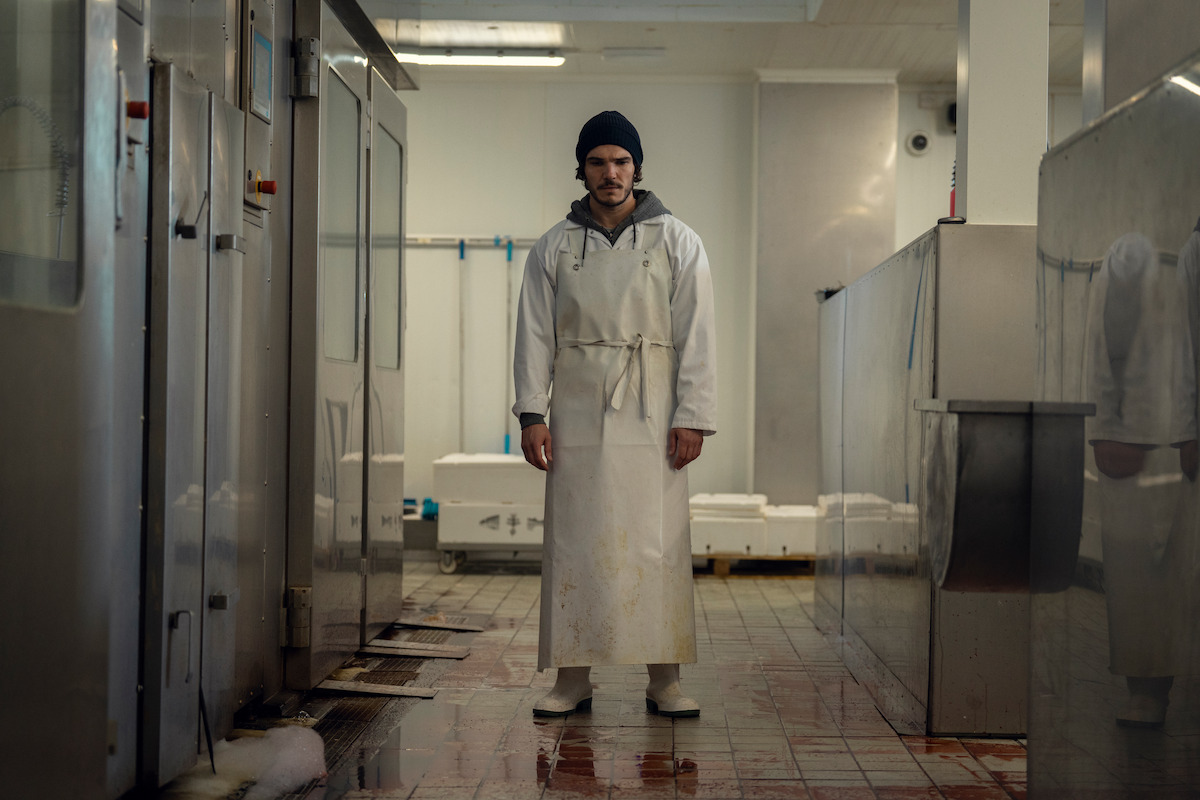 A man in a white apron and beanie stands in the center of a clean, industrial fish processing facility with tiled floors and stainless steel equipment.